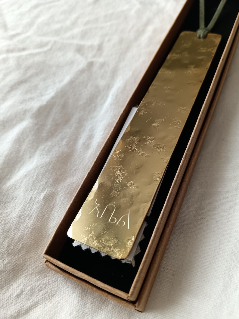 Peut inclure: Un marque-page rectangulaire en m&eacute;tal dor&eacute; avec une texture de surface martel&eacute;e et r&eacute;fl&eacute;chissante. Le marque-page pr&eacute;sente des caract&egrave;res grav&eacute;s en bas et est attach&eacute; &agrave; un fin cordon vert, pr&eacute;sent&eacute; dans une bo&icirc;te cadeau rectangulaire en carton brun.