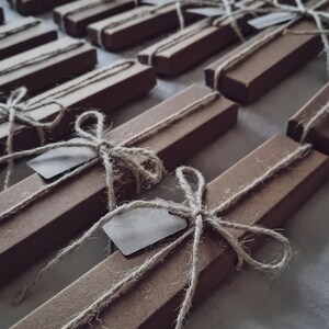 Peut inclure: Une collection de bo&icirc;tes rectangulaires en carton brun dispos&eacute;es en rang&eacute;es. Chaque bo&icirc;te est entour&eacute;e d'une fine ficelle de couleur claire nou&eacute;e en boucle au centre. Une petite &eacute;tiquette en papier rectangulaire vierge est gliss&eacute;e sous la ficelle.
