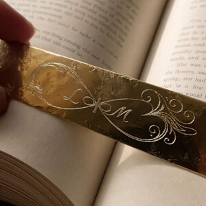 Peut inclure: Un marque-page rectangulaire en m&eacute;tal dor&eacute; orn&eacute; d'un symbole de l'infini grav&eacute;. Le centre de la boucle de l'infini contient les lettres L et M en &eacute;criture cursive, entour&eacute;es de volutes d&eacute;coratives complexes aux deux extr&eacute;mit&eacute;s.