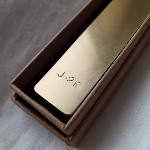 Könnte beinhalten: Ein rechteckiges Lesezeichen aus goldfarbenem Metall mit den eingravierten Buchstaben J und K, die durch ein kleines Herzsymbol getrennt sind. Das Lesezeichen liegt in einer einfachen, offenen braunen Pappschachtel mit glatter Oberfläche.