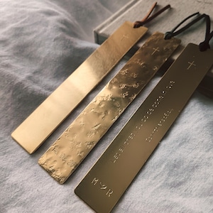 May include: Three brass bookmarks with leather cords. One bookmark has a smooth surface, another has a textured surface, and the third has engraved text. Each bookmark features a small cross at the top.