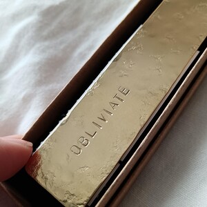 Peut inclure: Une plaque rectangulaire en m&eacute;tal dor&eacute; avec une finition de surface textur&eacute;e et martel&eacute;e. Le mot OBLIVIATE est grav&eacute; en lettres capitales au centre de la plaque. La plaque est log&eacute;e dans une bo&icirc;te en carton brun aux bords visibles.