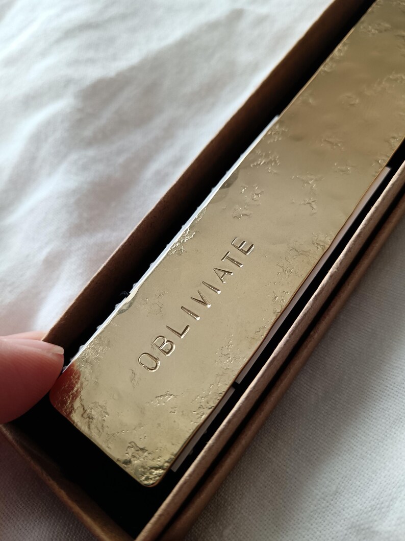 Peut inclure: Une plaque rectangulaire en m&eacute;tal dor&eacute; avec une finition de surface textur&eacute;e et martel&eacute;e. Le mot OBLIVIATE est grav&eacute; en lettres capitales au centre de la plaque. La plaque est log&eacute;e dans une bo&icirc;te en carton brun aux bords visibles.