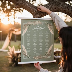 Op de afbeelding: Een saliegroene plattegrond met witte houten staven aan de boven- en onderkant. De kaart heeft een bloemenontwerp en de woorden "The Seating Chart". Het geeft de tafelnummers en gastnamen weer. De kaart wordt door een persoon omhoog gehouden.