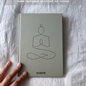 Op de afbeelding: Een grijs A5 yoga-dagboek met gelinieerde pagina's. De omslag heeft een lijntekening van een persoon in de sukhasana yoga-houding. De naam "ROBYN" staat op de omslag gedrukt.