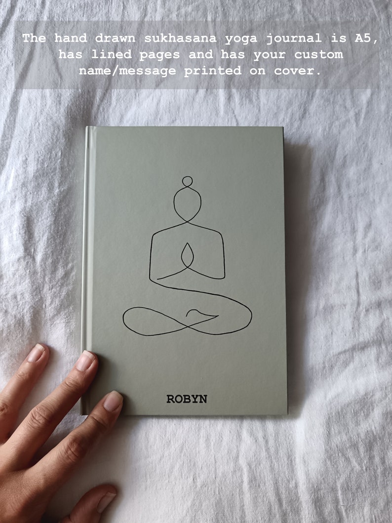Op de afbeelding: Een grijs A5 yoga-dagboek met gelinieerde pagina's. De omslag heeft een lijntekening van een persoon in de sukhasana yoga-houding. De naam "ROBYN" staat op de omslag gedrukt.