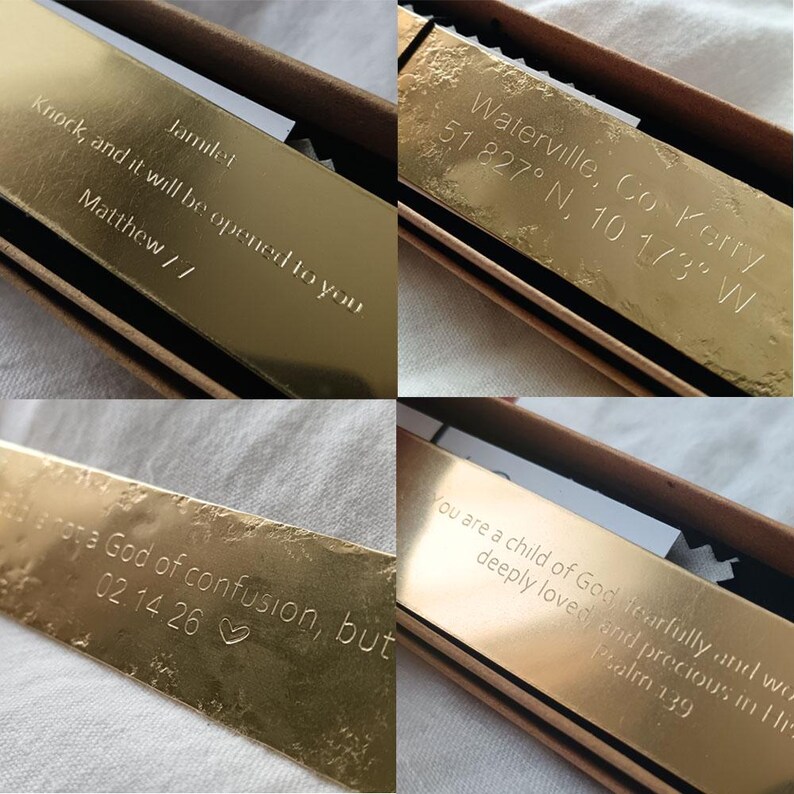 May include: A collection of four rectangular gold-toned metal bookmarks, each featuring custom laser-engraved text. The engravings include religious verses, geographic coordinates for Waterville, Co Kerry, names, and dates, all presented in a brown box.