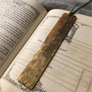 May include: A textured brass bookmark with a cross at the top and the numbers "14 09 26" engraved at the bottom. The bookmark is attached to a green cord and rests inside an open book.