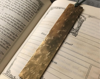 May include: A rectangular brass bookmark with a hammered, textured surface. It features a small engraved cross near the top and the date 14 09 26 stamped at the bottom. A dark green leather cord is threaded through a hole at the top edge.