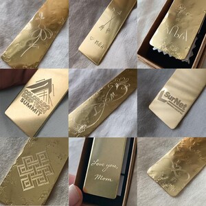 Peut inclure: Une collection de marque-pages rectangulaires en m&eacute;tal dor&eacute; pr&eacute;sentant divers motifs grav&eacute;s au laser, notamment une fleur, des fl&egrave;ches crois&eacute;es avec des initiales, le texte Colorado Deep Tech Summit, SurNet, Love you, Mom et des motifs g&eacute;om&eacute;triques.