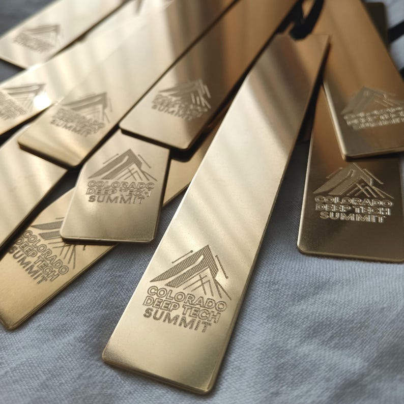 Peut inclure: Une collection de marque-pages rectangulaires en m&eacute;tal dor&eacute;, chacun pr&eacute;sentant un motif de montagne grav&eacute; et le texte "COLORADO DEEP TECH SUMMIT" centr&eacute; pr&egrave;s du bas. Les marque-pages ont une finition lisse et r&eacute;fl&eacute;chissante avec des coins arrondis.