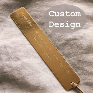 Puede incluir: Un marcapáginas de metal dorado con texto grabado y una borla de cuero. El marcapáginas tiene las palabras "Custom Design" en un óvalo gris. El texto grabado dice: "Las palabras pertenecen a todos los lugares donde se hacen humanos." - Rikki Bader Greenberg.