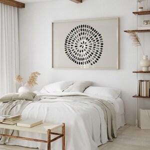 May include: A white bed with a white duvet cover and a beige throw blanket. The bed is in a bedroom with a white wall and a wooden beam ceiling. There is a black and white abstract print hanging on the wall above the bed.