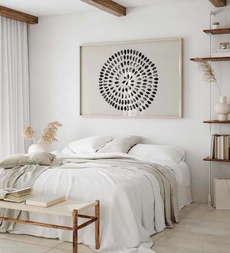 May include: A white bed with a white duvet cover and a beige throw blanket. The bed is in a bedroom with a white wall and a wooden beam ceiling. There is a black and white abstract print hanging on the wall above the bed.