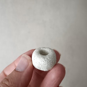 Puede incluir: Un pequeño objeto en forma de rosquilla, de color gris claro, con una superficie texturizada y un agujero central. El objeto se sostiene en una mano sobre un fondo neutro. El artículo parece estar hecho de piedra o un material similar.