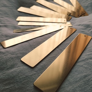 Puede incluir: Una colección de marcadores rectangulares de metal dorado dispuestos sobre una superficie texturizada de color gris azulado. Cada marcador es un rectángulo largo y delgado con un pequeño agujero en un extremo.