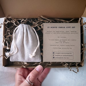 Op de afbeelding: Een 20 minuten durende kaars cadeau set in een kartonnen doos. De set bevat mini bijenwaskaarsen, een keramische kaarsenhouder en een katoenen trekkoordtas. De tekst op de kaart luidt: "Perfect voor meditatie, yoga, lezen, gebed of andere mindfulness praktijken."