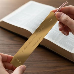 May include: A gold-colored metal bookmark with the text "Let all that you do be done in love" and "Corinthians 16:14". The bookmark has a cross symbol and a pink cord. The initials "M ♡ R" are at the bottom.