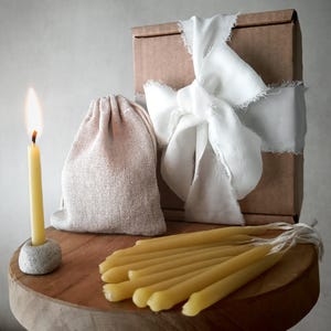 May include: A lit yellow candle in a stone holder, alongside a bundle of similar candles and a drawstring bag on a wooden surface. A brown box tied with a white ribbon is in the background. The scene suggests a natural, warm aesthetic.