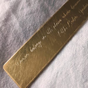 Peut inclure: Un marque-page rectangulaire en m&eacute;tal dor&eacute; avec une citation grav&eacute;e en &eacute;criture cursive qui dit, Women belong in all places where decisions are being made, suivie du nom Ruth Bader Ginsburg, avec une surface lisse et r&eacute;fl&eacute;chissante.