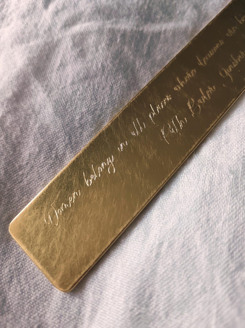 Peut inclure: Un marque-page rectangulaire en m&eacute;tal dor&eacute; avec une citation grav&eacute;e en &eacute;criture cursive qui dit, Women belong in all places where decisions are being made, suivie du nom Ruth Bader Ginsburg, avec une surface lisse et r&eacute;fl&eacute;chissante.