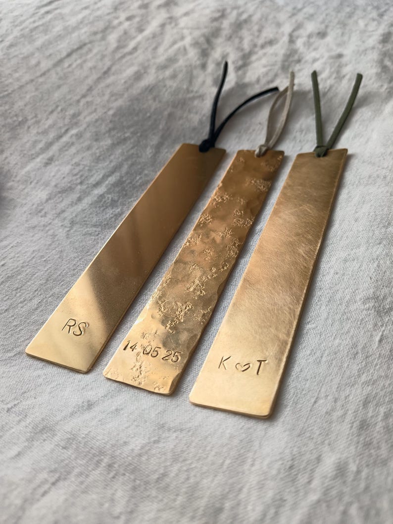 Peut inclure: Trois marque-pages rectangulaires en laiton aux coins arrondis avec des cordons. Le premier est lisse avec les lettres RS, le deuxi&egrave;me a une texture martel&eacute;e avec la date 14.05.25, et le troisi&egrave;me a une finition bross&eacute;e avec les lettres K et T s&eacute;par&eacute;es par un c&oelig;ur.