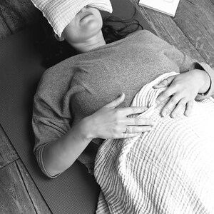 May include: A person lying on a yoga mat with a pillow over their eyes. They are wearing a grey shirt and have a blanket over their lower body. The person is practicing mindfulness and relaxation.