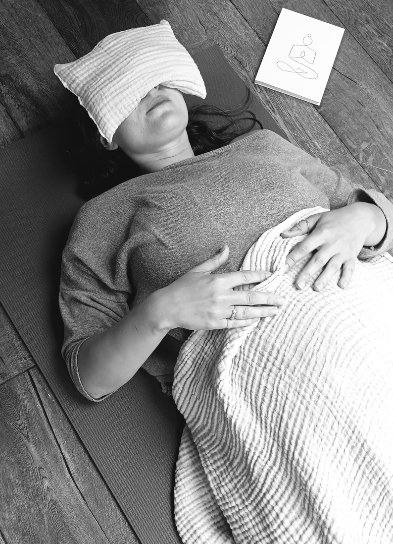 May include: A person lying on a yoga mat with a pillow over their eyes. They are wearing a grey shirt and have a blanket over their lower body. The person is practicing mindfulness and relaxation.
