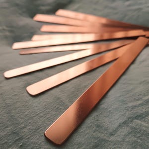 Puede incluir: Varias tiras metálicas de color cobre dispuestas sobre una tela turquesa. Las tiras, rectangulares con extremos redondeados, reflejan la luz. Primer plano que destaca la textura y el color metálico.