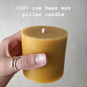 Op de afbeelding: Een close-up van een 100% pure bijenwas-pilaarvormige kaars. De kaars is lichtgeel van kleur en wordt vastgehouden door een hand. De kaars heeft een witte lont.