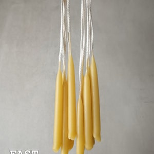May include: A bunch of thin, yellow beeswax candles with white wicks hanging from a hand. The candles are about 10 inches long.  FAST SHIPPING is written in white text on a gray background.