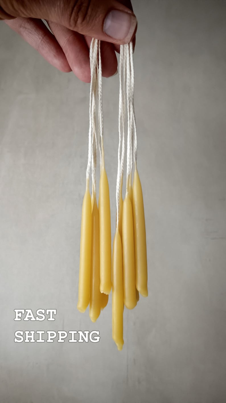 May include: A bunch of thin, yellow beeswax candles with white wicks hanging from a hand. The candles are about 10 inches long.  FAST SHIPPING is written in white text on a gray background.