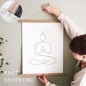 Pode incluir: Um cartaz vertical branco com um desenho de linhas pretas de uma pessoa numa posição de meditação sentada. O cartaz é mantido por calhas magnéticas de madeira clara no topo e na base. Uma inserção mostra opções de calhas brancas, de carvalho e pretas.