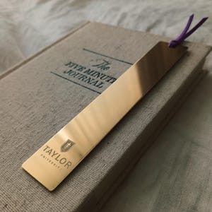 May include: A rectangular gold-toned metal bookmark with a reflective surface, featuring the Taylor University logo and text engraved at the bottom. A thin purple ribbon is tied through a hole at the top of the bookmark, which rests on a beige book.
