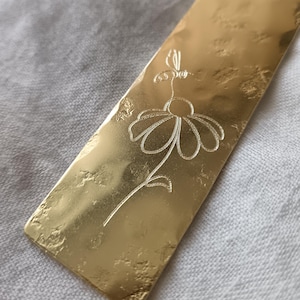 Puede incluir: Un marcapáginas rectangular de metal dorado con una superficie martillada y texturizada. El marcapáginas presenta una ilustración grabada de líneas finas de una margarita con una pequeña abeja sobrevolando el centro de los pétalos.