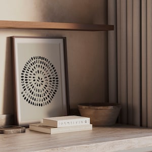 May include: A framed print of a black and white circular design with a stack of two books on a light brown countertop. The top book has the title "100% LIVING" and the bottom book has the title "Yves De Braban - Works from the studio".