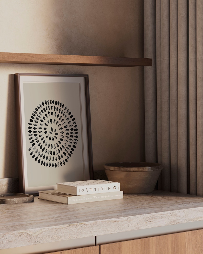 May include: A framed print of a black and white circular design with a stack of two books on a light brown countertop. The top book has the title "100% LIVING" and the bottom book has the title "Yves De Braban - Works from the studio".