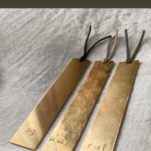Peut inclure: Trois marque-pages rectangulaires en laiton avec des cordons en cuir. Le premier est lisse avec les lettres RS, le deuxi&egrave;me a une texture martel&eacute;e avec la date 14 05 25, et le troisi&egrave;me a une finition bross&eacute;e avec les lettres K et T et un c&oelig;ur.