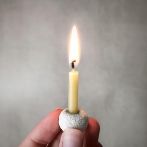 Puede incluir: Una vela amarilla delgada y encendida con una llama brillante, sostenida en un pequeño soporte blanco texturizado. La vela se encuentra sobre un fondo gris suave, creando un ambiente cálido y acogedor.