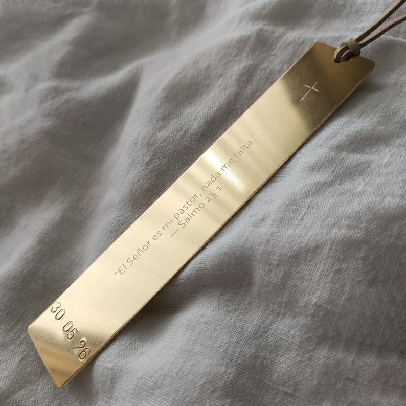 May include: A rectangular gold-colored metal bookmark with a smooth, reflective finish. It features an engraved cross at the top, the Spanish text "El Se&ntilde;or es mi pastor, nada me falta - Salmo 23:1" in the center, and the date "30 05 26" at the bottom.