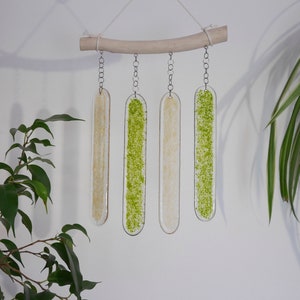 May include: A decorative wind chime featuring four clear, elongated glass panels suspended from a natural wooden branch. Two panels have a beige textured design, while the other two have a vibrant green speckled pattern. The chime hangs against a white wall.