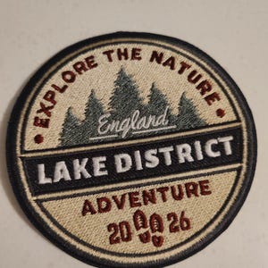 May include: Circular embroidered patch with the text "EXPLORE THE NATURE" and "LAKE DISTRICT ADVENTURE" in white and red lettering. The patch features a forest scene and the word "England". The patch has a black border.