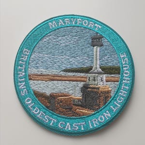 May include: Circular embroidered patch depicting Maryport's oldest cast iron lighthouse. The teal border contains the text "MARYPORT" and "BRITAIN'S OLDEST CAST IRON LIGHTHOUSE." The image shows a lighthouse, sea, and land.