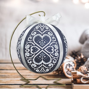 May include: A blue and white Christmas bauble with a heart pattern is displayed on a gold metal stand. The bauble is surrounded by a festive setting with a small artificial Christmas tree, cinnamon sticks, and a wooden box.