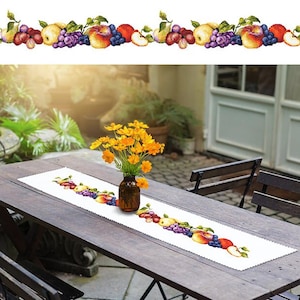 Könnte beinhalten: Ein weißer Tischläufer mit einem bunten, gestickten Früchte-Design. Der Läufer liegt auf einem Holztisch mit zwei Stühlen. Ein Strauß gelber Blumen steht in einer Vase auf dem Tisch.