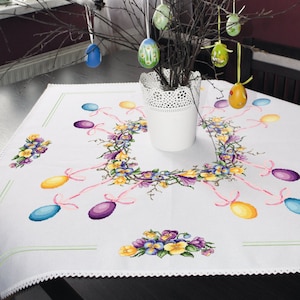 Puede incluir: Mantel blanco con diseño floral bordado y huevos de Pascua de colores pastel. El mantel tiene un delicado borde de encaje y está decorado con una corona de flores y huevos de Pascua de colores pastel.