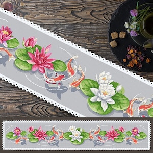 Op de afbeelding: Een grijze en witte tafelrunner met een witte kantzoom heeft een herhalend patroon van roze en witte waterlelies, groene waterleliebladeren en oranje vissen.