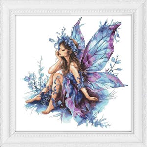 May include: A framed cross-stitch artwork depicts a seated fairy with blue and purple wings. The fairy wears a blue dress and a floral crown. The artwork is set against a white background and framed in a white, ornate frame.