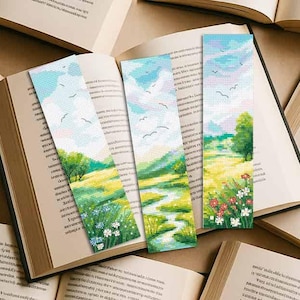 Könnte beinhalten: Drei Lesezeichen mit Landschaftsmotiven im Kreuzstich-Design liegen in einem offenen Buch. Die Lesezeichen zeigen einen Himmel mit Wolken, Bäumen, Feldern und Blumen. Die Lesezeichen sind in Blau-, Grün-, Gelb- und Weißtönen gehalten.