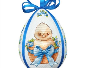 Può includere: Un uovo di Pasqua bianco con un nastro blu e un piccolo pulcino giallo all'interno di un cestino con fiori blu. Il pulcino indossa un fiocco blu.